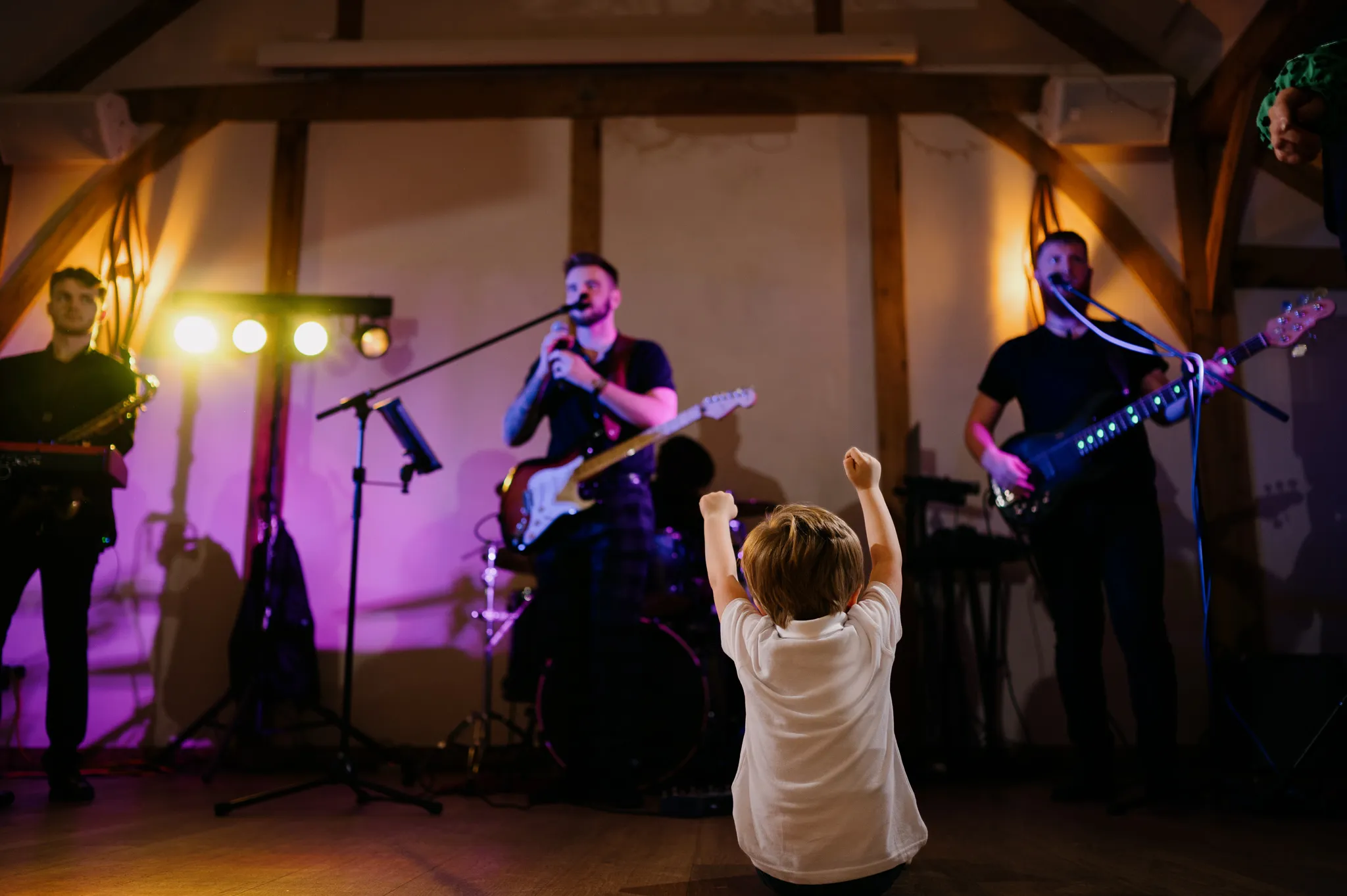 Young child in a white shirt raising arms facing a live band performing on stage with guitars and a saxophone under warm lighting.